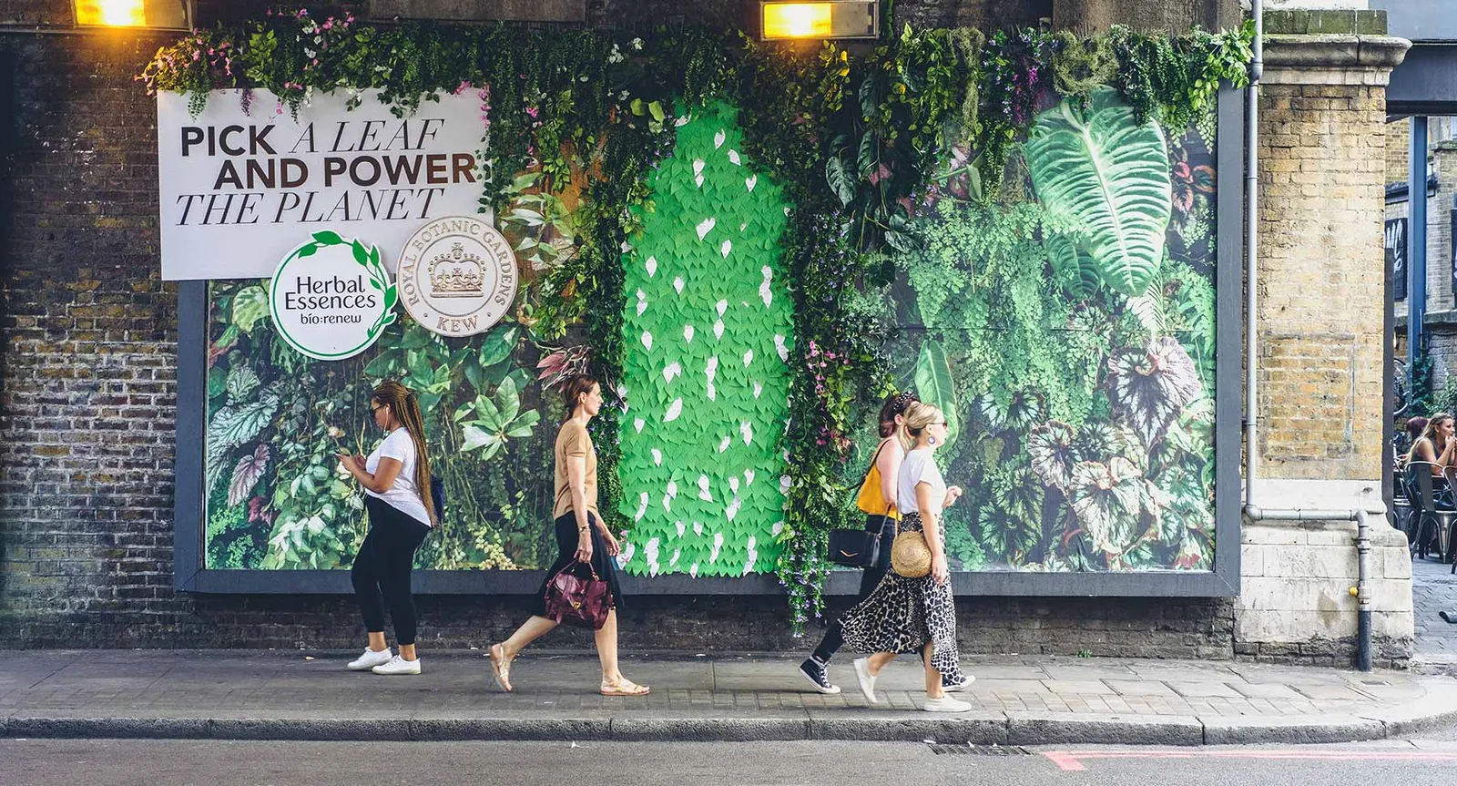 Herbal Essences billboard headed “Pick a leaf and power the planet”, covered in green paper leaves and foliage, with pedestrians walking past