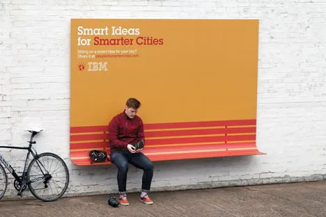 Orange IBM “Smart Ideas for Smarter Cities” billboard whose lower edge folds out into a bench, with a man sitting on it