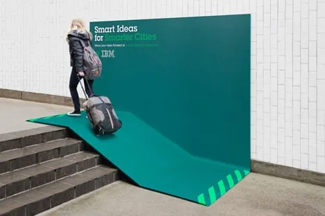 Green IBM “Smart Ideas for Smarter Cities” billboard bent over a few steps to form a ramp, with a woman pulling a suitcase up it