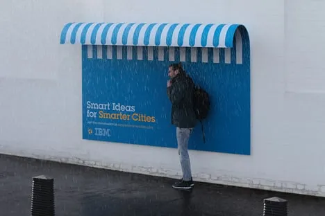 Blue IBM “Smart Ideas for Smarter Cities” billboard curving out at the top into a striped awning that keeps the rain off a man standing beneath it