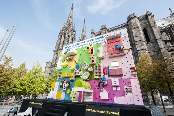 IKEA Clermont-Ferrand climbing wall: furniture fixed to a vertical wall like an apartment turned on its side, with climbers in harnesses, in front of the cathedral