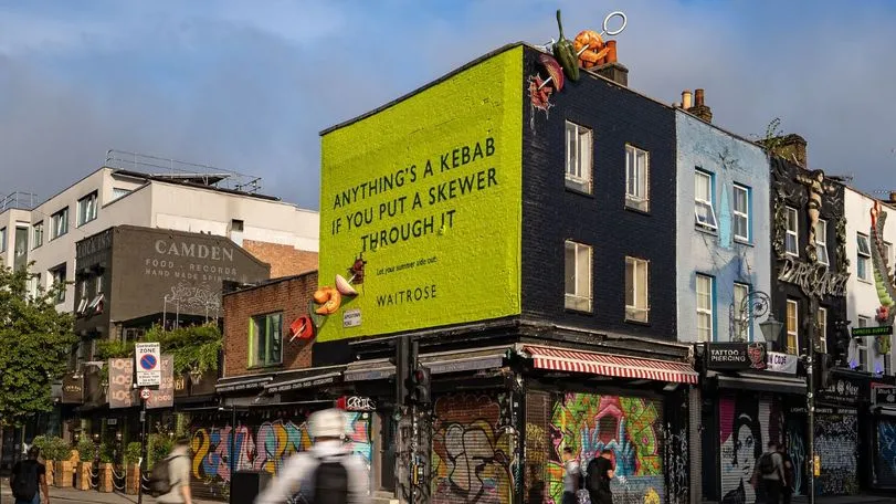 Lime-green painted end wall of a building in Camden with the Waitrose line “Anything’s a kebab if you put a skewer through it” and giant skewered food breaking through the brickwork