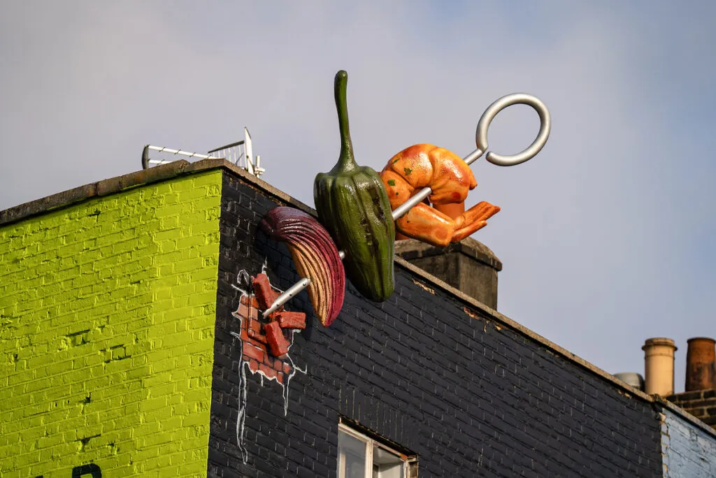 Close-up of the giant skewer with a red onion, a green pepper and a prawn emerging from the corner of a brick building